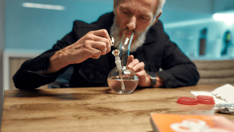 Senior man lighting cannabis in the bowl of glass water pipe or bong in the kitchen.