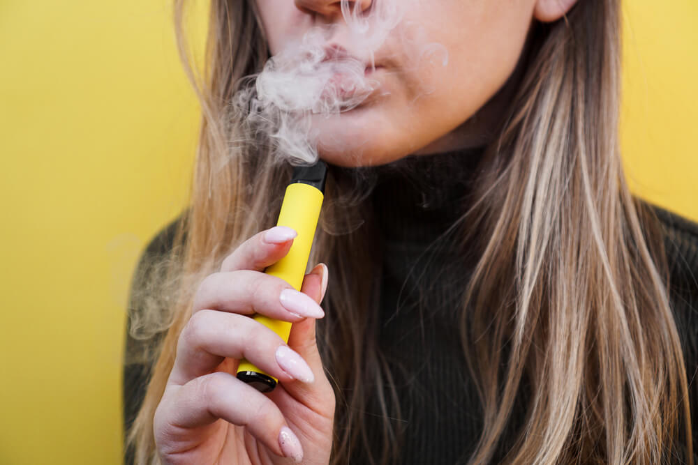 A young girl smokes disposable electronic cigarette. Bright yellow background 