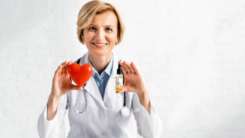 cheerful and mature doctor holding bottle with cbd lettering and red heart in clinic
