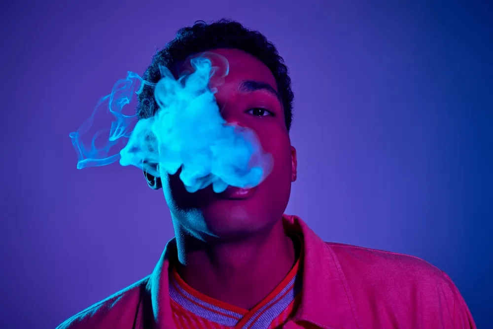 young african american man exhaling smoke against blue background with purple lighting